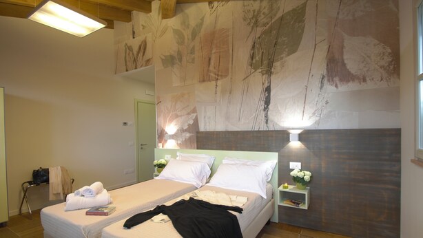 Habitación de hotel con dos camas, una mesita de noche con lámpara, un estante en la pared con flores y un mural abstracto grande.