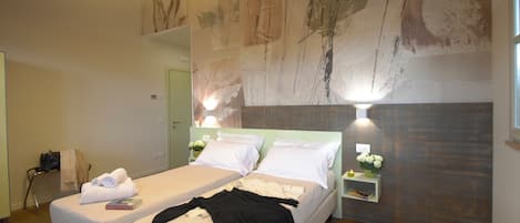A hotel room with two beds, a nightstand with a lamp, a wall-mounted shelf with flowers, and a large abstract mural on the wall.