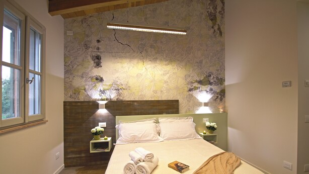 Una cama bien hecha con toallas enrolladas, dos lámparas de noche, una mesita de noche con un florero y un cabecero de madera.