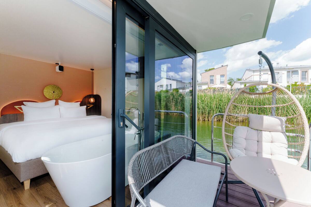 A modern bedroom with a large bed, a glass door leading to a balcony with a wicker chair and a small table.
