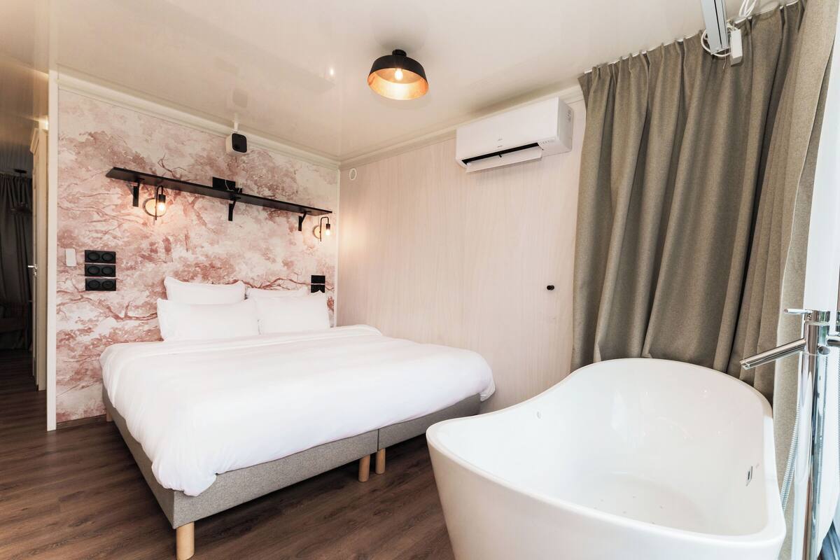 A bedroom with a large bed, a built-in shelf, an air conditioning unit, and a freestanding bathtub.
