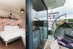 A modern bedroom with a bed, a glass door leading to a balcony with wicker chairs and a view of boats and buildings. - L'Escale Royale L'Isle Adam Port Oise (L'Isle-Adam)