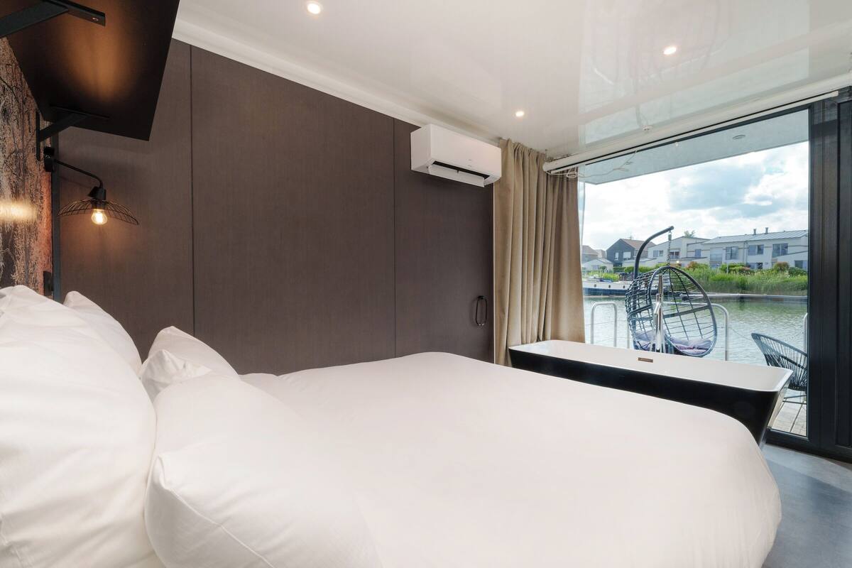 A modern hotel room with a large bed, a small table, and a view of a canal with boats.