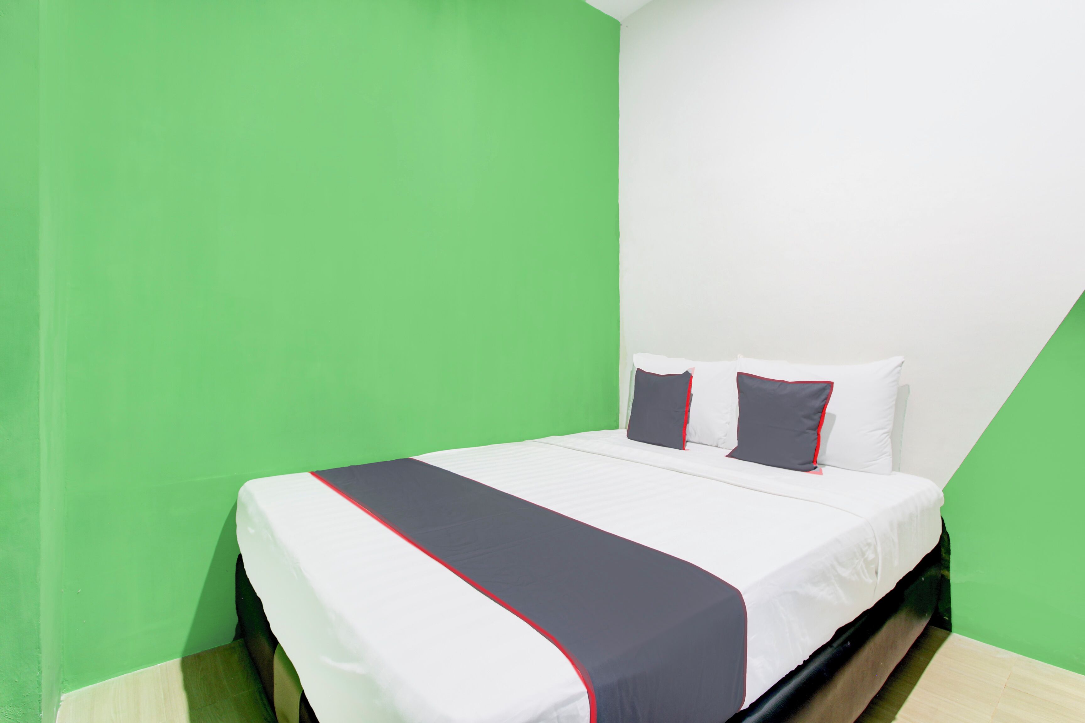 Una cama individual con una colcha blanca y roja, dos almohadas y un cabecero negro en una habitación con paredes verdes y techo blanco.