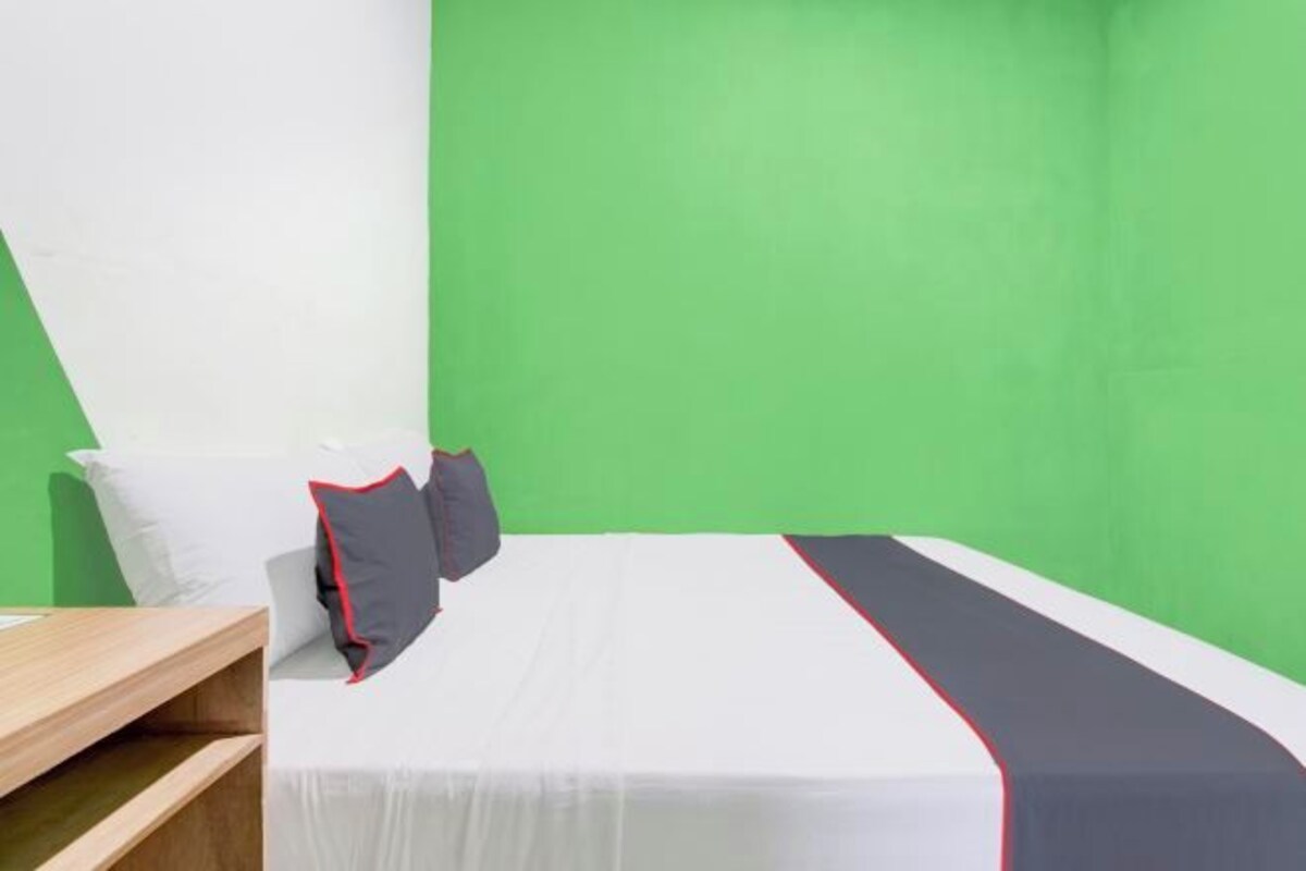 A single bed with white bedding and a red and black accent, a wooden nightstand, and a green wall.
