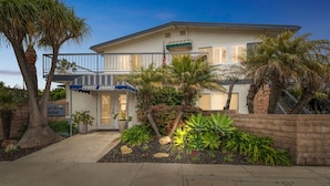 A two-story building with a balcony, a sign for Sunset Shores, and a well-maintained garden with various plants and a palm tree. - Beautiful 1 Bed Beach Condo #224 1 Bedroom Condo by Sunset Shores Condominiums (Carpinteria)