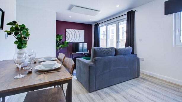 A modern living room with a grey sofa, a dining table with plates and glasses, and a TV in the background.
