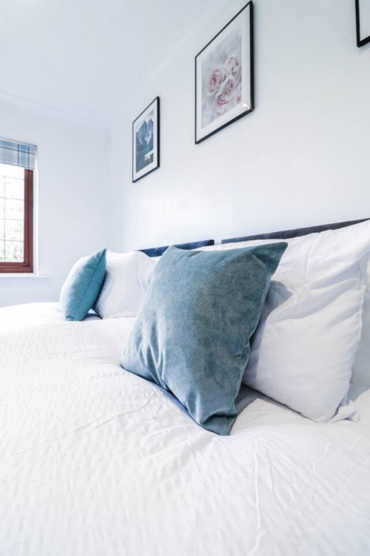 Un lit bien fait, avec une literie blanche et bleue, une fenêtre avec des stores et deux tableaux encadrés accrochés au mur.