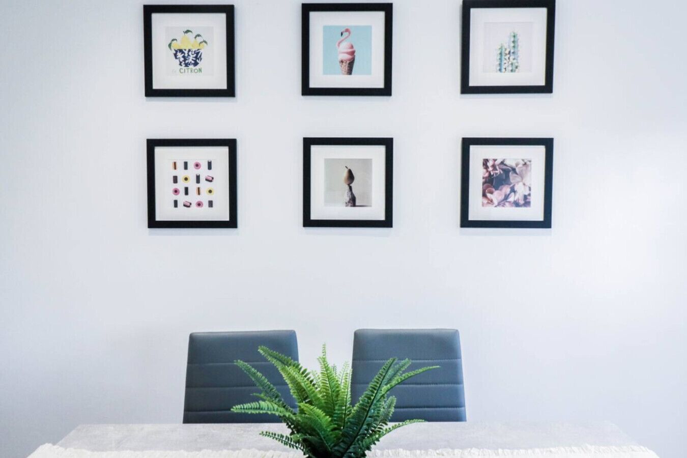 A room with a white wall, six framed artworks, two grey chairs, and a green plant.