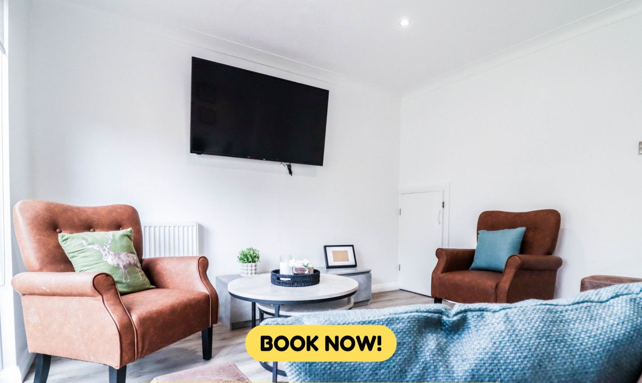 A modern living room with two leather armchairs, a round coffee table, and a flat-screen TV mounted on the wall. Amenities include free WiFi, parking, self-check-in, cooking, and laundry services.
