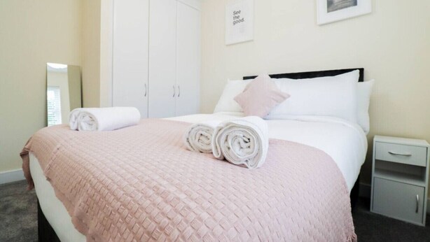 Una cama bien tendida con toallas enrolladas, un armario blanco, una mesita de noche pequeña y un cuadro enmarcado en la pared.