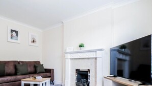Una sala de estar moderna con un sofá de cuero marrón, una mesa de centro blanca, una chimenea y un televisor sobre un soporte blanco. Los servicios incluyen WiFi gratuito, estacionamiento, acceso independiente, cocina y lavandería.