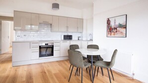 A modern kitchen with white cabinetry, a built-in oven, and a microwave. There is a dining area with a round table and four chairs. - 2BR Seaside Sanctuary in St Leonards on Sea Flat 5 (Hastings)