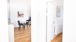 A modern kitchen with white cabinets, a dining area with a round table and chairs, and a hallway with a white door. - 2BR Seaside Sanctuary in St Leonards on Sea Flat 5 (Hastings)