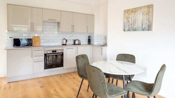 Une cuisine moderne avec des meubles blancs, un four à micro-ondes, un grille-pain et une bouilloire. On y trouve une table à manger avec quatre chaises et un tableau accroché au mur.