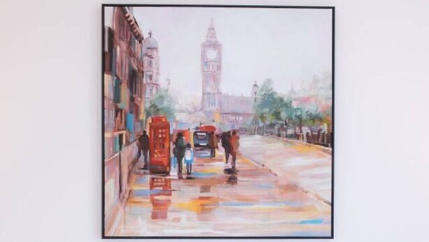 A painting of a street scene with people and a red telephone booth, mounted on a white wall above a gray headboard.