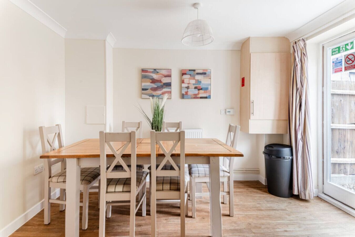 A dining area with a wooden table and chairs, two abstract paintings on the wall, a radiator, a trash bin, and a window with curtains.
