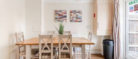 A dining area with a wooden table and chairs, two abstract paintings on the wall, a radiator, a trash bin, and a window with curtains.