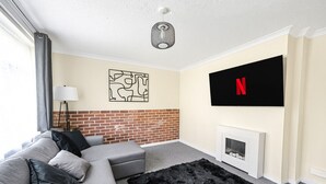 A living room with a grey sofa, black and grey cushions, a black furry rug, a fireplace, a TV mounted on the wall, and a brick accent wall. - The Ipswich Snug Spacious 3BR Retreat FREE Parking (Ipswich)