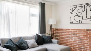 A living room with a grey sectional sofa, a floor lamp, and a brick wall. - The Ipswich Snug Spacious 3BR Retreat FREE Parking (Ipswich)
