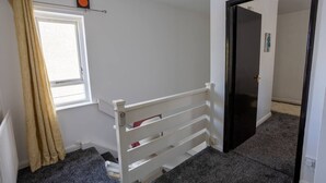 Una habitación con una cama blanca, una ventana con cortinas color beige, un radiador y una puerta que da a otra habitación.