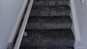 Una escalera con peldaños alfombrados y barandillas blancas, que lleva a un rellano con un radiador y una silla.