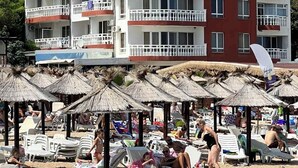 A hotel with multiple balconies, a red and white facade, and a beach area with sun loungers and umbrellas. - Hotel NOY (Varna)