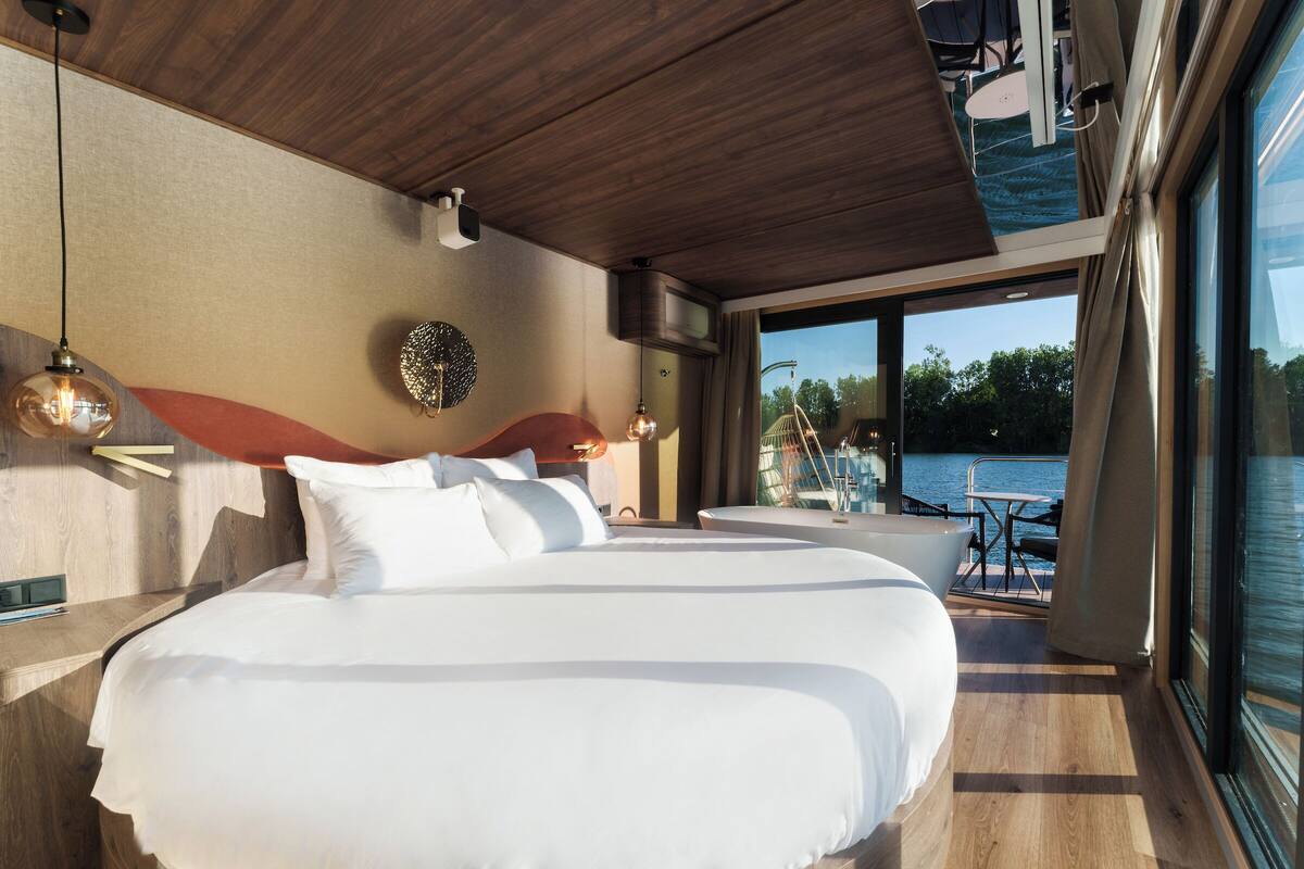 Un dormitorio moderno con una cama grande, techo de madera y vista a un cuerpo de agua a través de amplias ventanas.