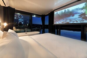 A hotel room with a bed, a large window, a projector screen displaying a waterfall, and a dining area with chairs and a table.