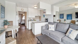 A modern kitchen with white cabinets, a central island, and a dining area with wicker furniture. - Beautiful 1 Bed Beach Balcony Condo #207 1 Bedroom Condo by RedAwning (Carpinteria)