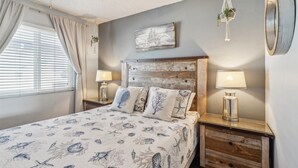 A bedroom with a bed, bedside tables, lamps, a ceiling fan, a window with curtains, and a wall-mounted plant. - Beautiful 1 Bed Beach Balcony Condo #207 1 Bedroom Condo by RedAwning (Carpinteria)
