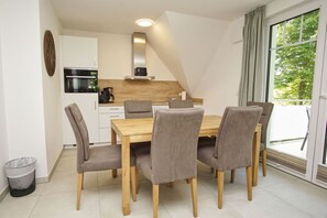 A modern kitchen with white cabinets, a wooden dining table, and six grey upholstered chairs. - Beachfront apartment with balcony, use of swimming pool and sauna - Rex Rugia apartment 17-5 - (Göhren)