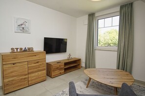 Uma sala de estar moderna com um suporte de TV de madeira, uma televisão de tela plana, uma mesa de madeira redonda e uma janela com cortinas.