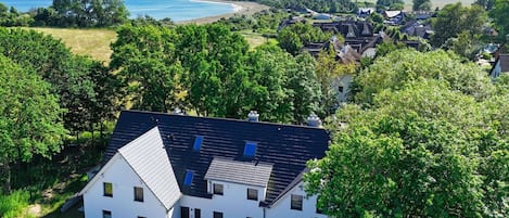 Une maison moderne à deux étages, dotée d’un toit en pente, entourée d’une végétation luxuriante et offrant une vue sur une plage et des collines lointaines.