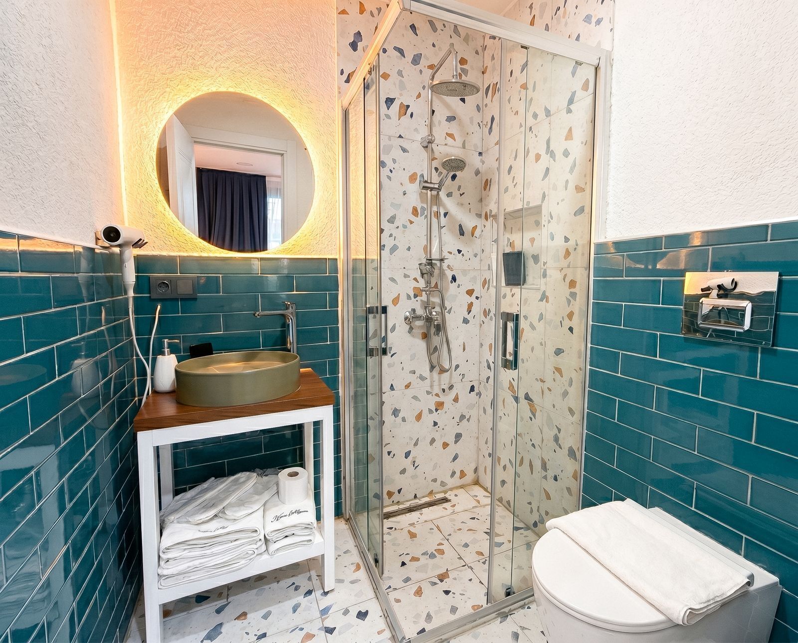 Une salle de bain dotée d’une douche à parois de verre, d’un lavabo avec un plan de travail en bois, d’un porte-serviettes et d’un miroir à cadre circulaire.