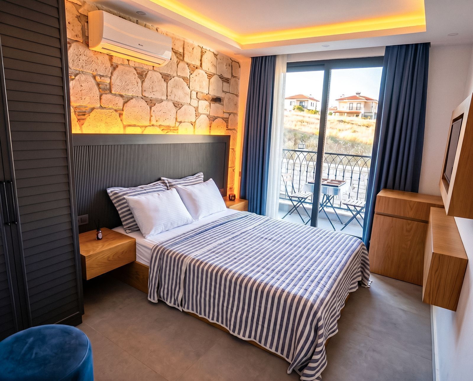 A bedroom with a stone wall, a bed with striped bedding, a nightstand, a wooden dresser, and a balcony with outdoor furniture.