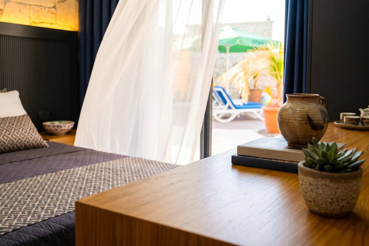 Ein Schlafzimmer mit einem Bett, einem Nachttisch mit Vase und Pflanze, einem Fenster mit Vorhängen und Blick auf einen Außenbereich mit Stuhl und Pflanze.