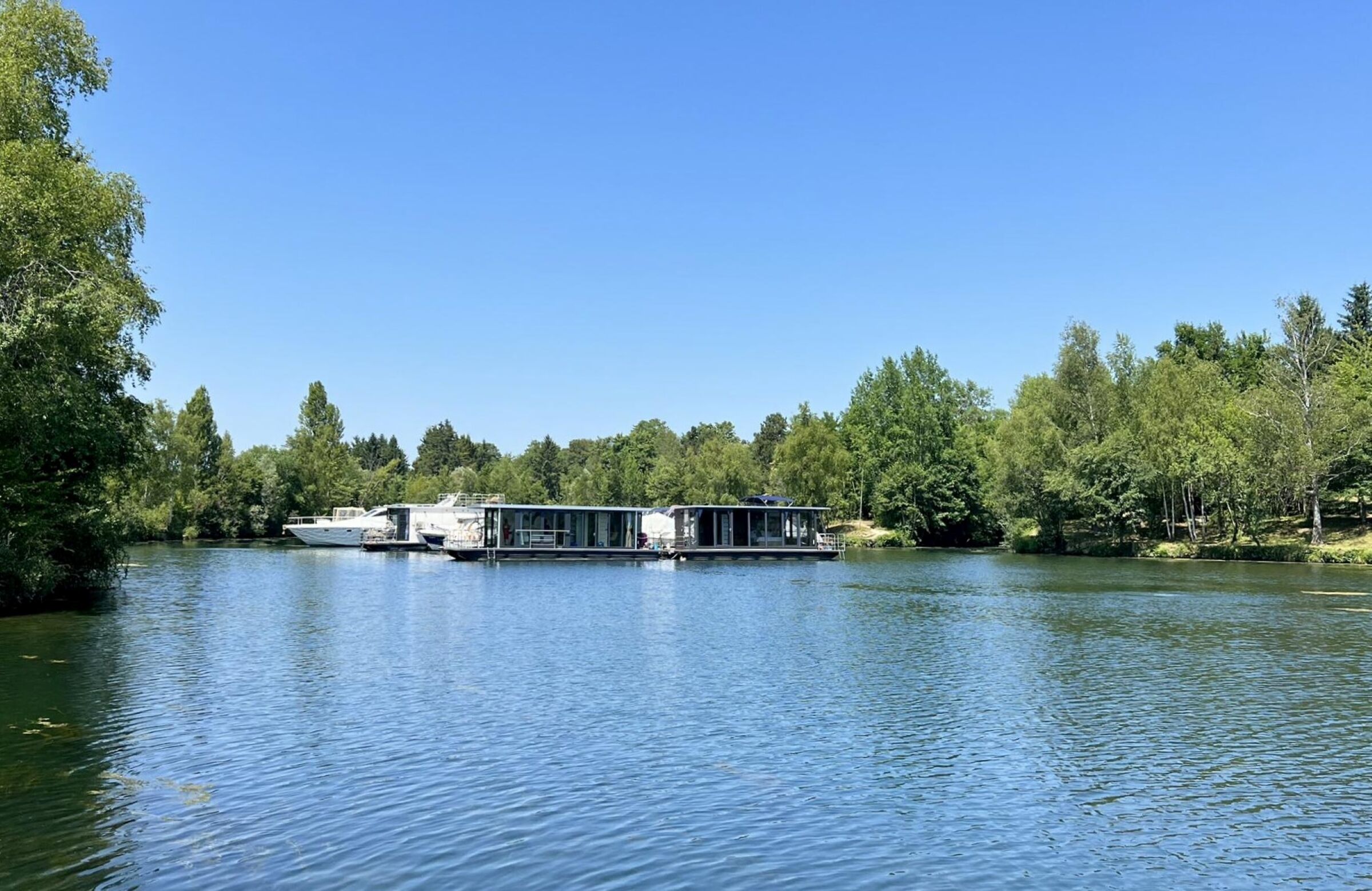 Uma casa flutuante atracada em um lago calmo, cercada por árvores densas.