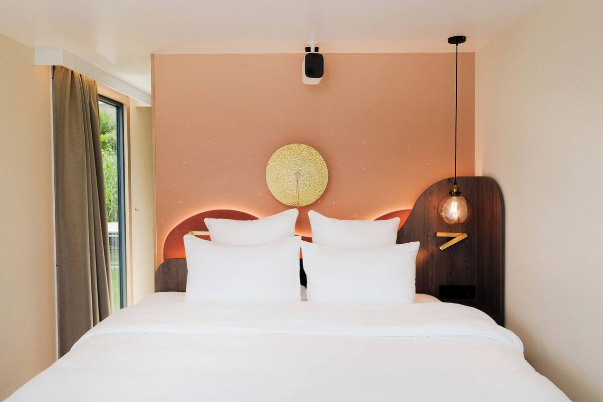 A modern hotel room with a large bed, a round wall decoration, a ceiling light, and a window with curtains.