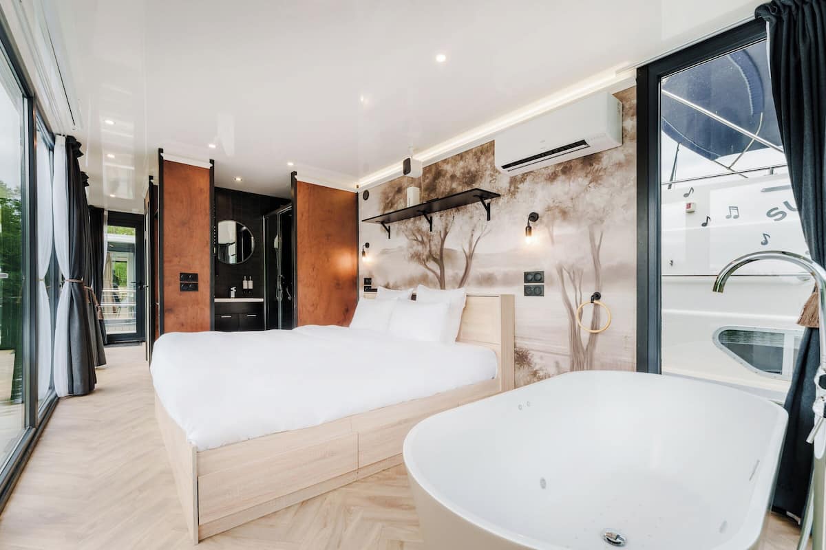 A modern bedroom with a large bed, a freestanding bathtub, and a view of the outdoors through large windows.