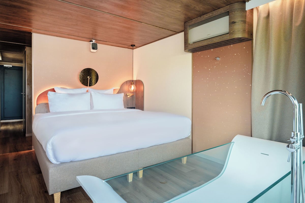 A modern hotel room with a large bed, a glass-enclosed bathtub, and a wooden ceiling.