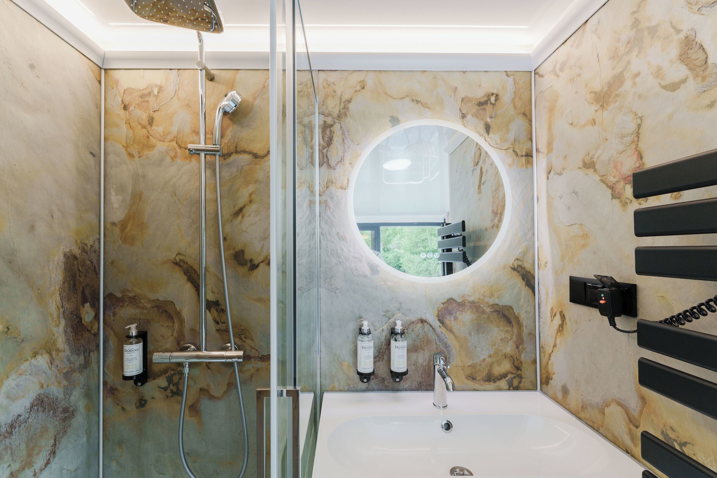 A bathroom with a marble wall, a round mirror, a shower, and a sink with two bottles of Dove soap.
