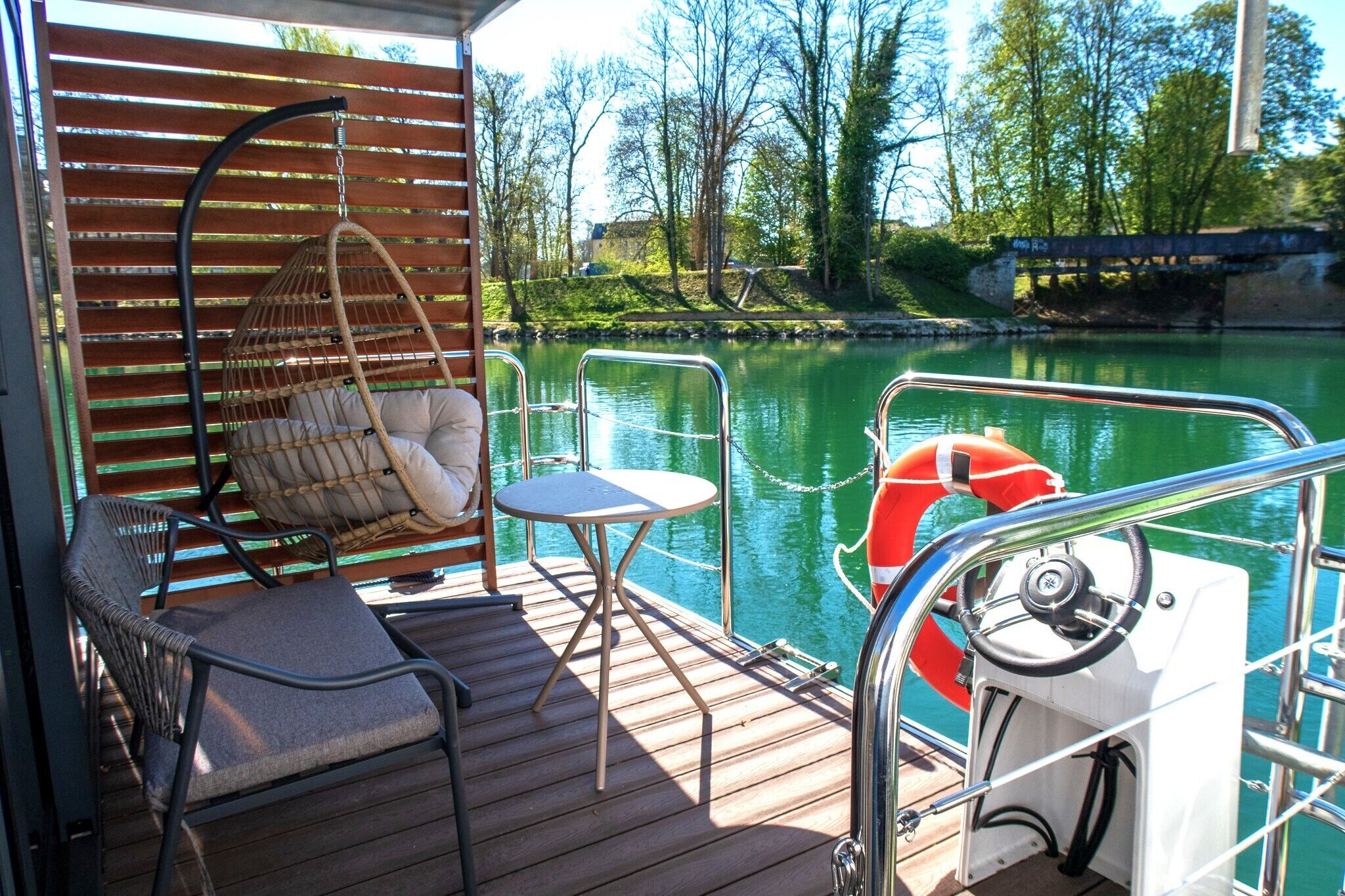 Une terrasse en bois avec un fauteuil suspendu, une petite table ronde et un volant de bateau.