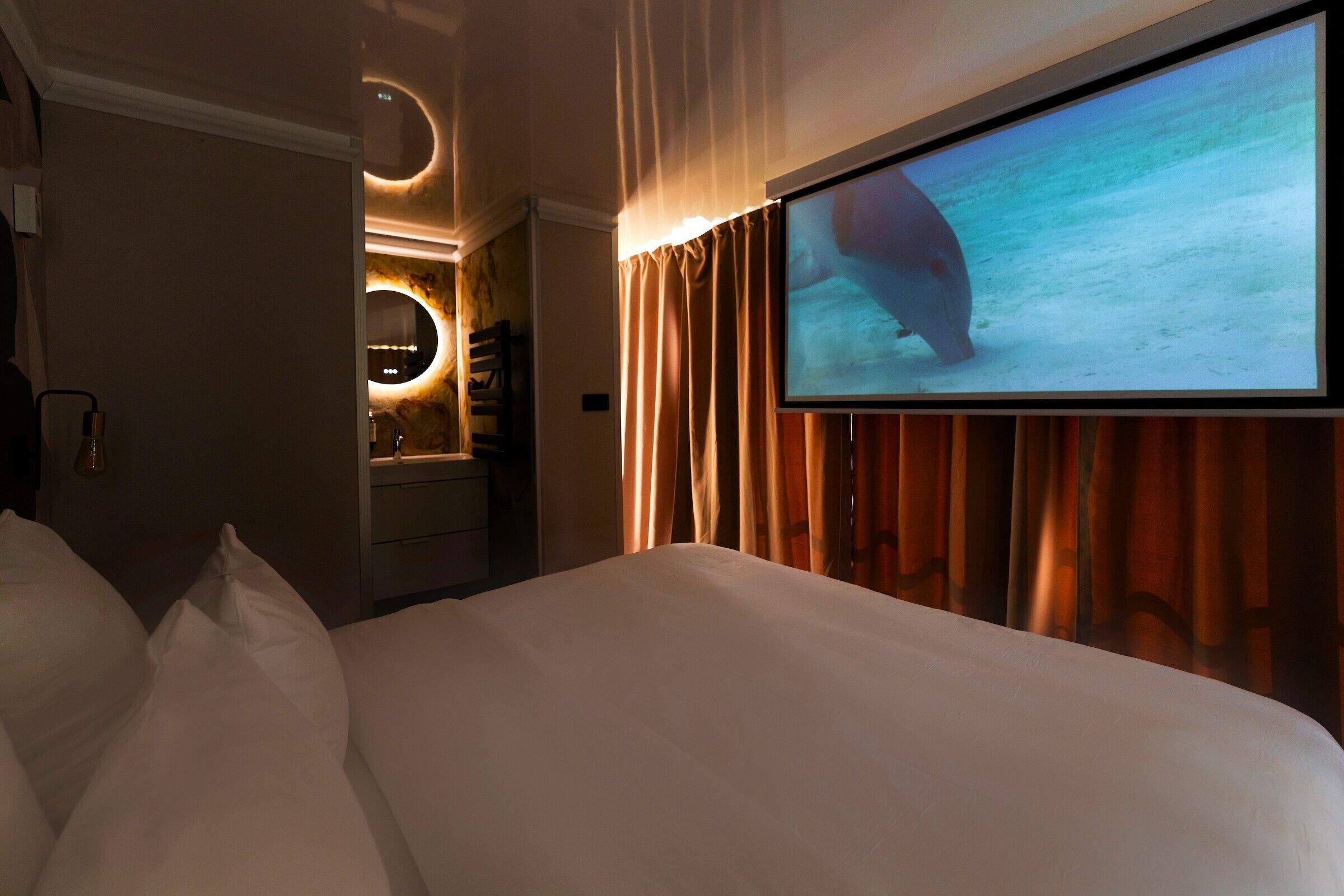 A hotel room with a bed, a TV screen displaying an underwater scene, a bathroom with a mirror, and curtains.