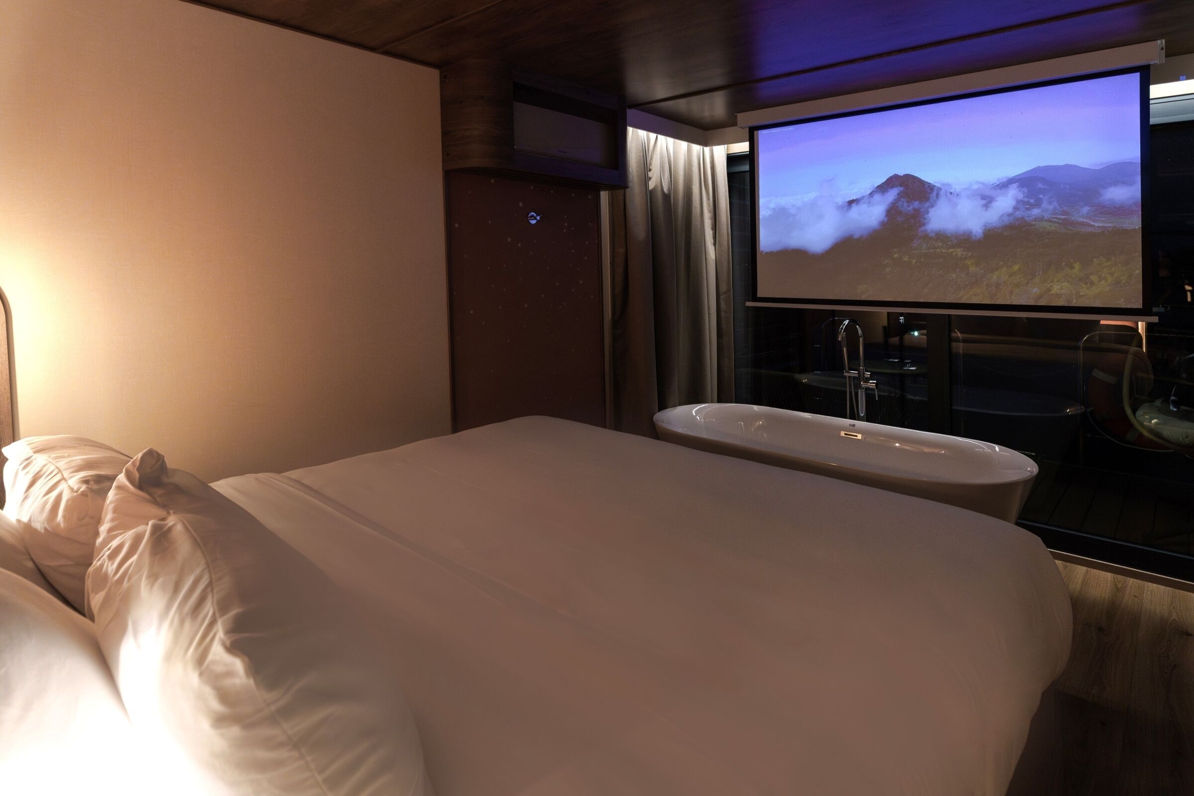 Ein Hotelzimmer mit Bett, großem Bildschirm, der eine Berglandschaft zeigt, Badezimmer mit Waschbecken und Duschbereich.