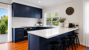 A modern kitchen with dark blue cabinets, a white island with bar stools, a built-in oven, and a large window with blinds. - Horizon Loft by Ready Set Stay (Cowes)