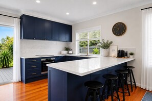 Une cuisine moderne avec des meubles de couleur bleu foncé, un îlot central blanc doté de tabourets, un four encastré et une grande fenêtre avec des stores.