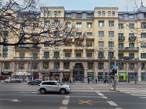 A multi-story building with a central entrance, storefronts on the ground floor, and a pedestrian crossing. - New luxury flat in the heart of Budapest (Budapest)