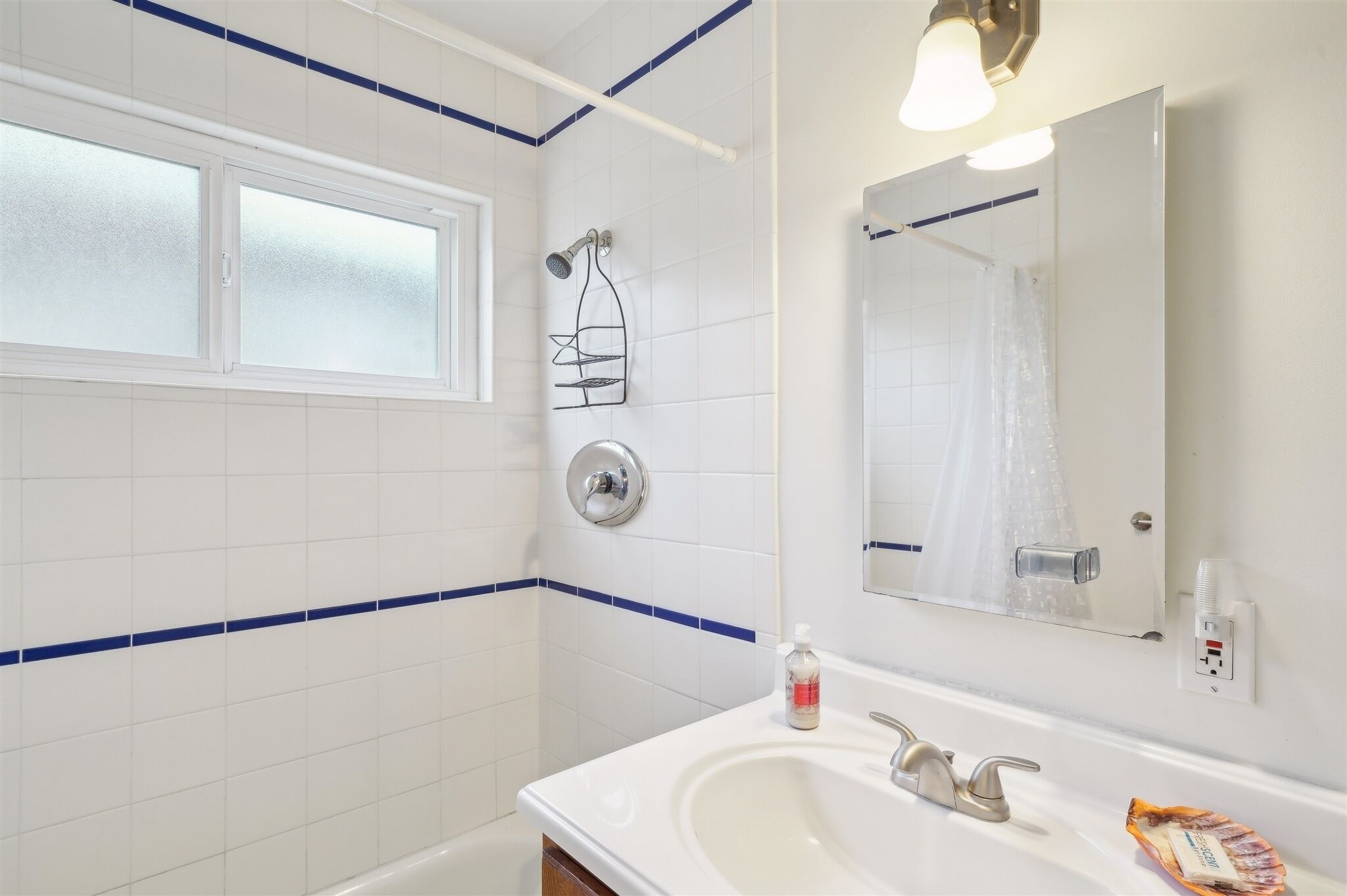 A bathroom with white tiles, a window, a shower, a sink, a mirror, and a towel rack.