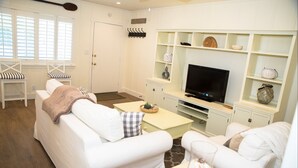 A living room with white furniture, a television, and shelves with decorative items. - 4980 Sandyland Road #216 2 Bedroom Condo by RedAwning (Carpinteria)